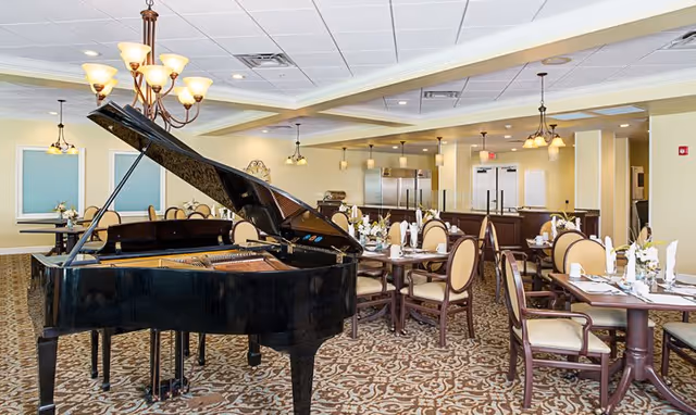 A spacious dining room in a senior living facility featuring a black grand piano in the foreground. The room has multiple round and rectangular tables set with white napkins, plates, and silverware. The chairs have cushioned seats and backs. The ceiling has recessed lighting and chandeliers, and the walls are painted light yellow with decorative accents. The floor is covered with a patterned carpet.