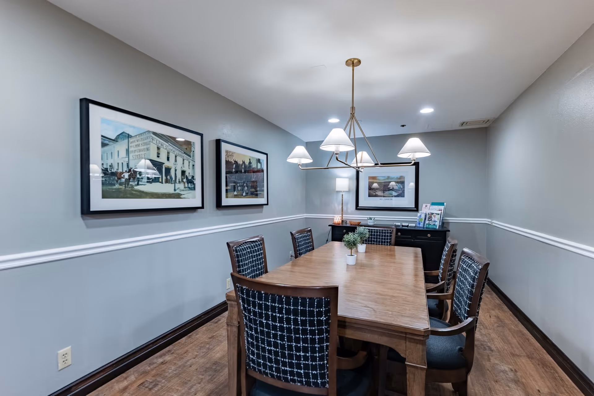 A dining room with a rectangular wooden table surrounded by six chairs with patterned upholstery. The room has light gray walls with white trim, three framed pictures on the walls, a modern chandelier with five lampshades hanging above the table, and a sideboard with a lamp and some brochures in the background.