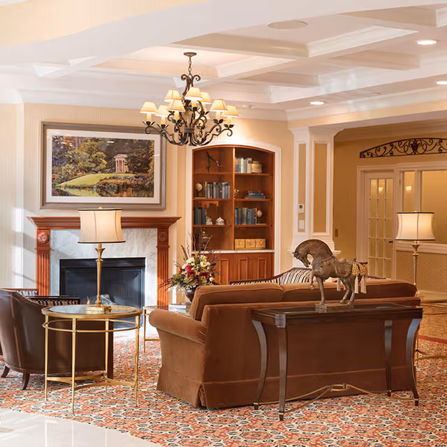 A cozy living room area in an assisted living facility featuring a brown sofa, two brown armchairs, a wooden console table with a horse sculpture, a round glass-top side table with a lamp, a fireplace with a framed landscape painting above it, a built-in wooden bookshelf filled with books and decorative items, and a chandelier hanging from a coffered ceiling.