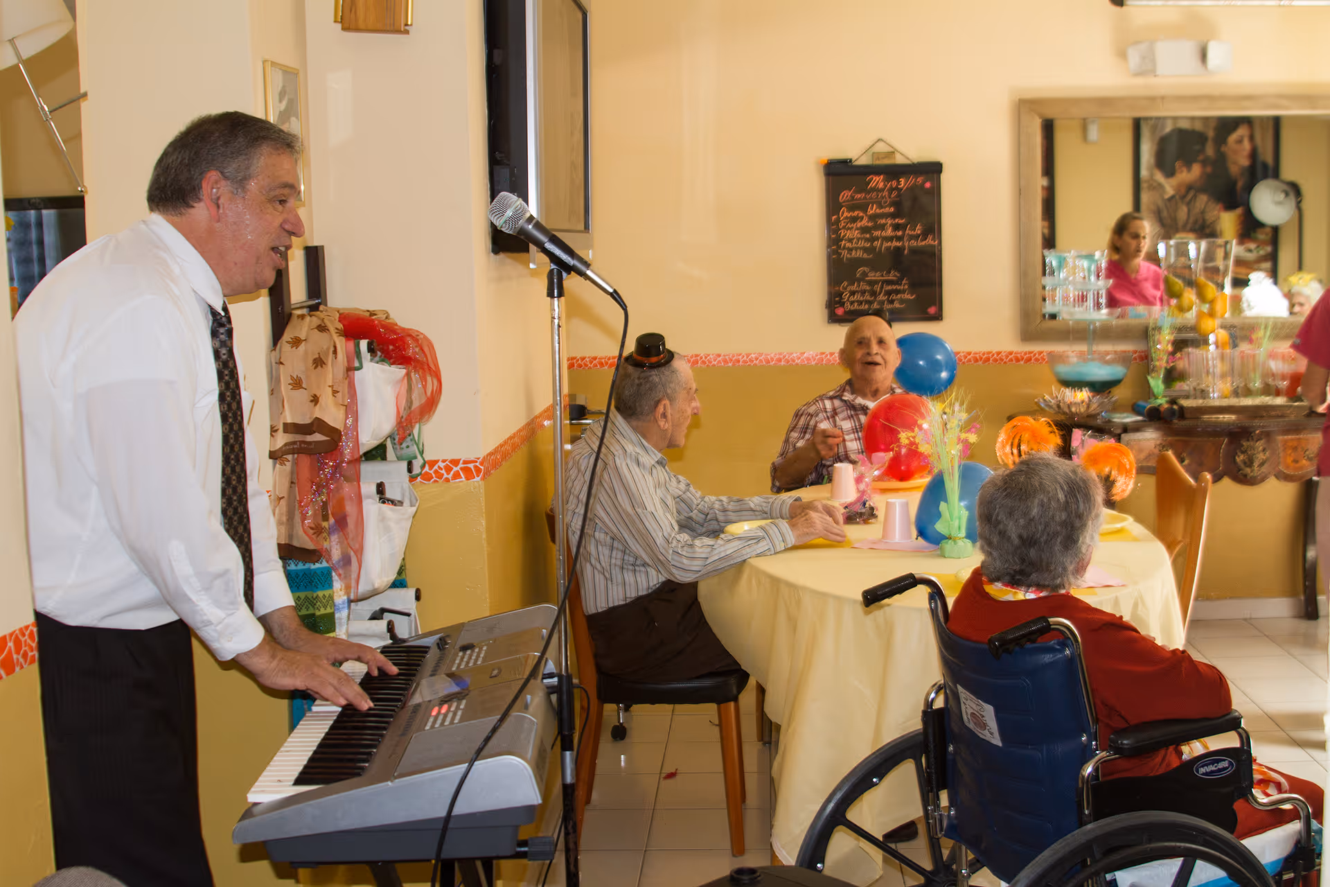 An elderly man plays a keyboard and sings into a microphone in a room with several elderly people seated around a table decorated with colorful balloons and flowers. One person is in a wheelchair, and the room has warm yellow walls with a mirror and a chalkboard menu on the wall.