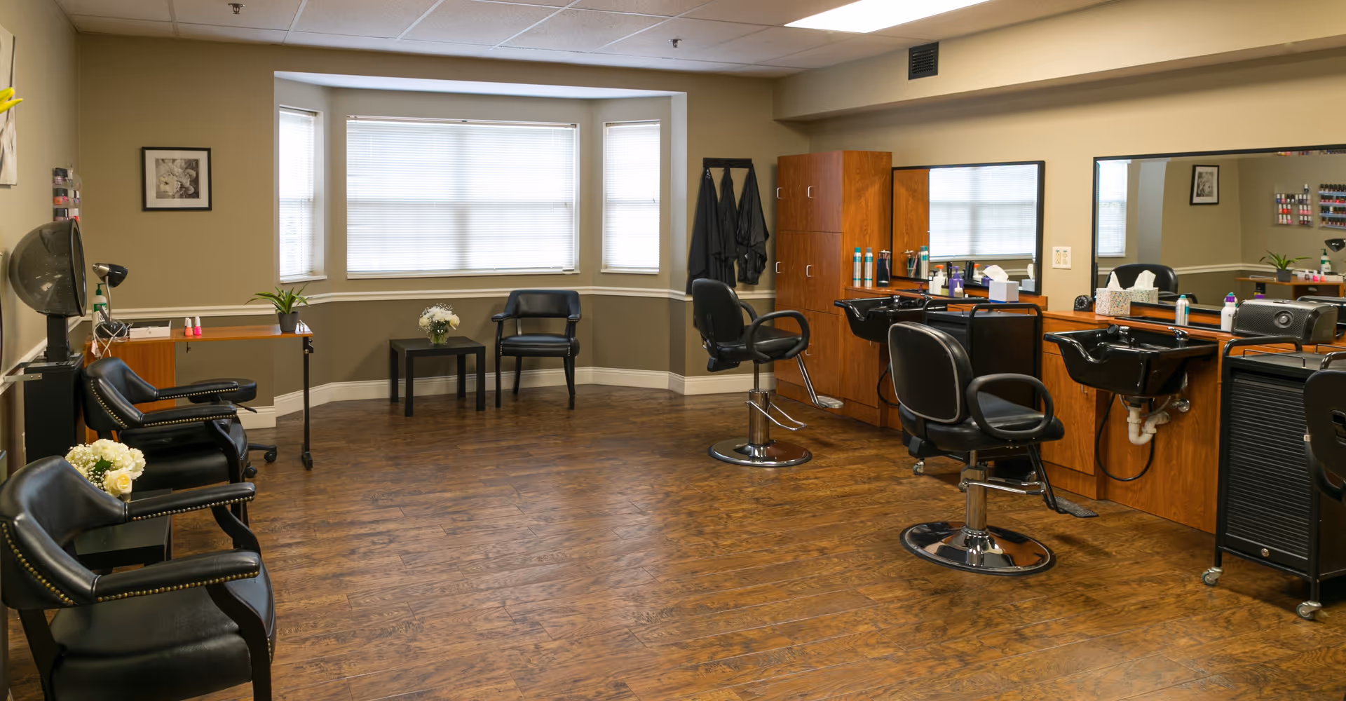 A clean and well-lit salon area in a senior living facility with wooden flooring, black salon chairs, hair washing sinks, large mirrors, and a window with blinds. There are also small tables with flowers and a hair dryer visible.