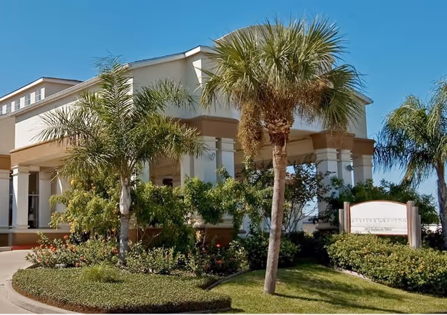 Exterior view of a senior living facility building with white walls and a beige trim, surrounded by palm trees and well-maintained landscaping under a clear blue sky. A sign is visible near the entrance.