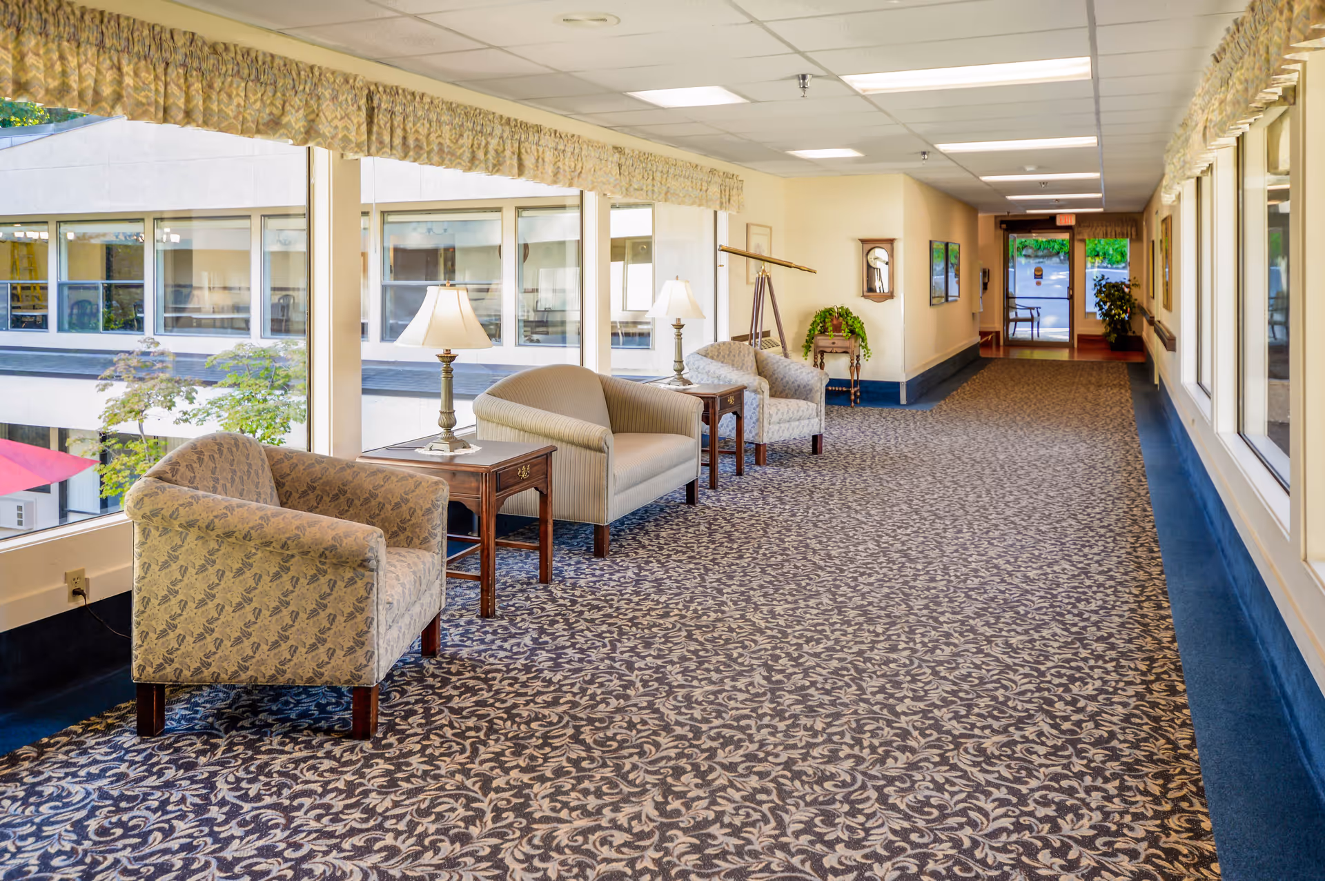 A bright and spacious hallway in an assisted living facility with large windows on one side, patterned carpet, and several upholstered chairs and small tables with lamps arranged along the wall. The hallway leads to a glass door with a view of greenery outside.