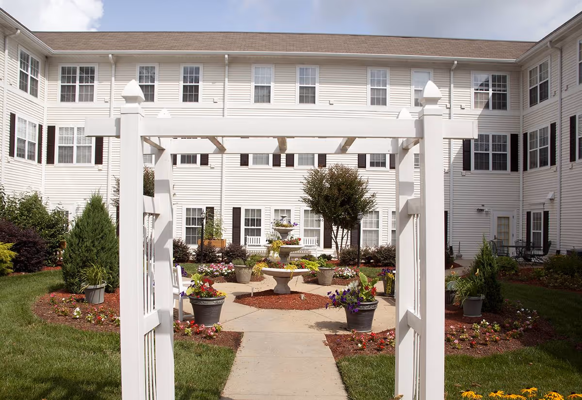 Outdoor courtyard area of Brighton Gardens of Raleigh featuring a white wooden pergola entrance, a central fountain surrounded by flower pots and landscaped garden beds, with a multi-story white building with many windows in the background.