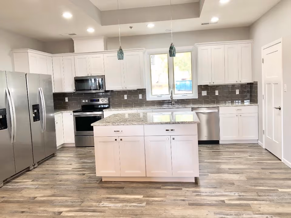 A modern kitchen with white cabinets, a large island with granite countertop, stainless steel appliances including a double-door refrigerator, oven, microwave, and dishwasher. The kitchen has a gray tile backsplash, wood-look flooring, two pendant lights hanging above the island, and a window above the sink letting in natural light.