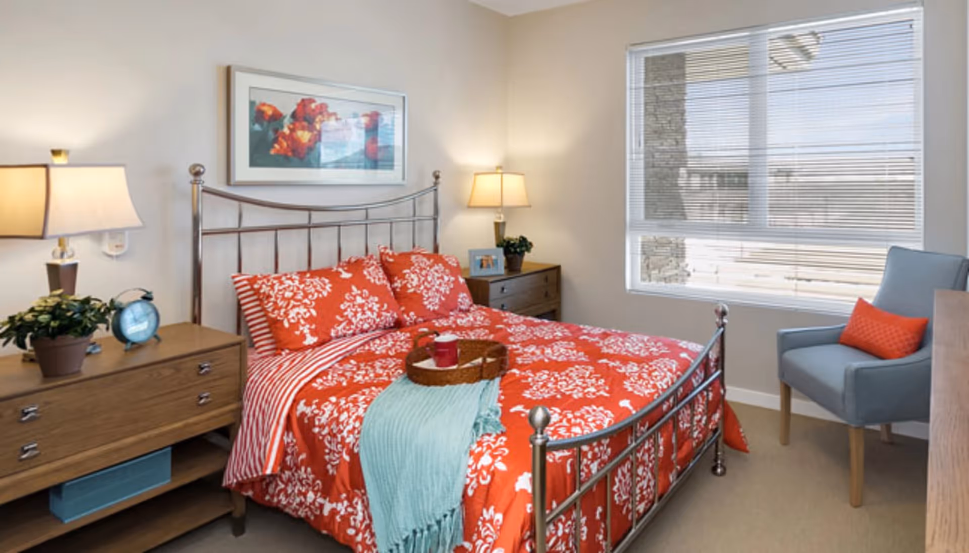 A bright bedroom with a metal frame bed dressed in red and white floral bedding and a light blue throw. There are two wooden nightstands on either side of the bed, each with a lamp and decorative items including a plant and a clock. A blue armchair with an orange pillow is positioned near a window with blinds, and a colorful framed artwork hangs above the bed.