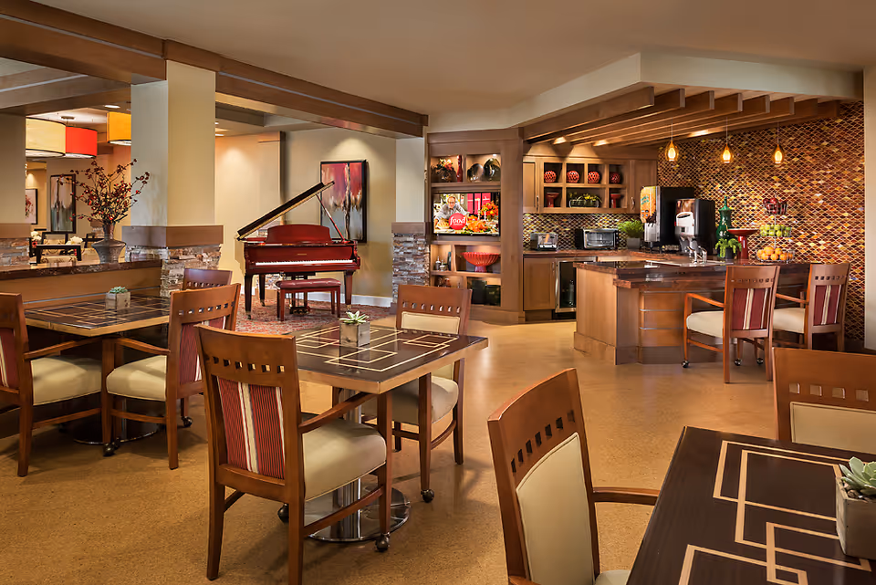 Interior view of a senior living facility common area featuring a dining space with wooden tables and chairs, a kitchen area with modern appliances and a mosaic tile backsplash, and a red grand piano in the background. The room is warmly lit with pendant lights and decorated with plants and artwork.