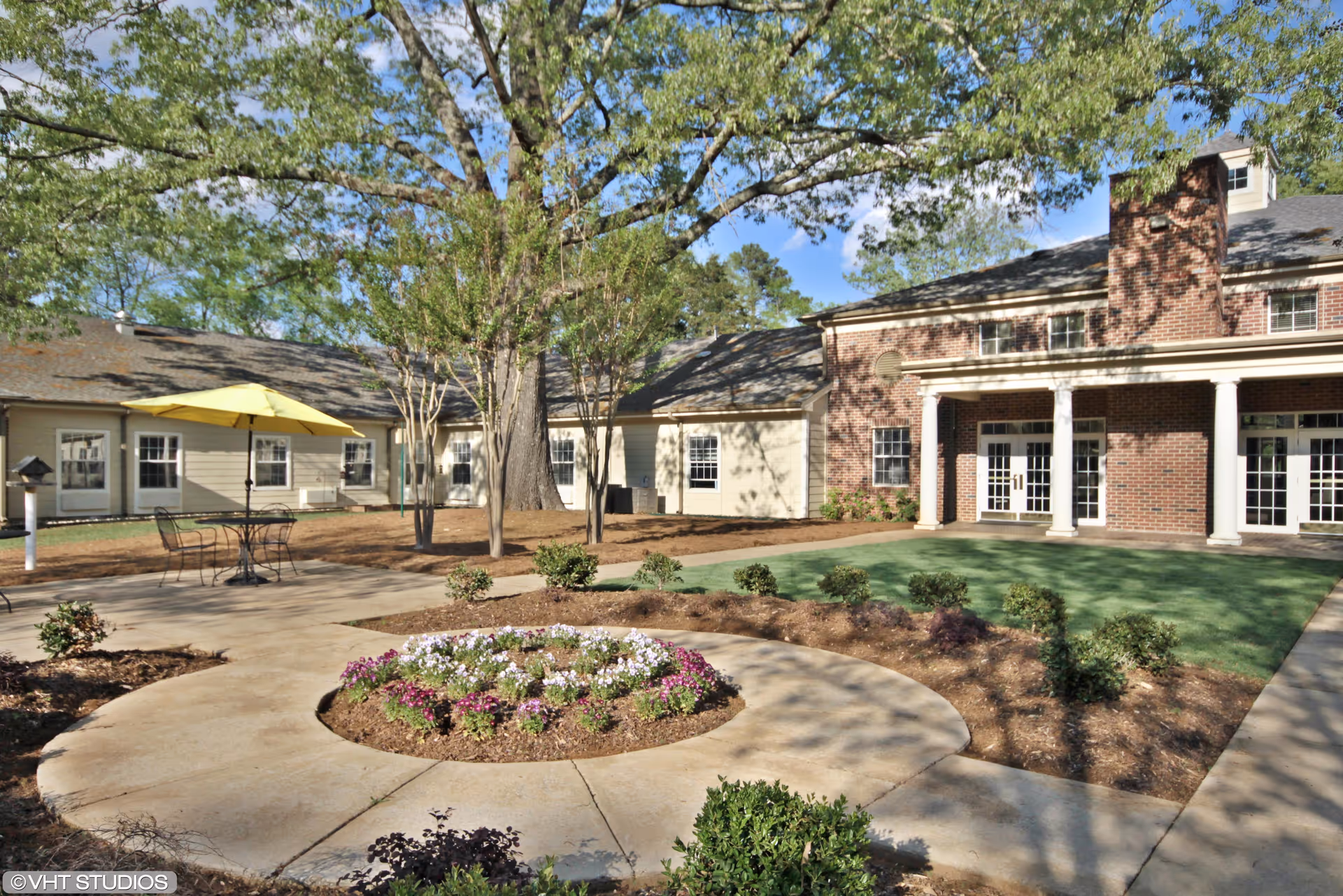Outdoor courtyard area at The Fairways at Grayson Valley featuring a circular flower bed with blooming flowers, a paved walkway, a table with a yellow umbrella, and surrounding buildings with large windows and a brick facade under a clear blue sky.
