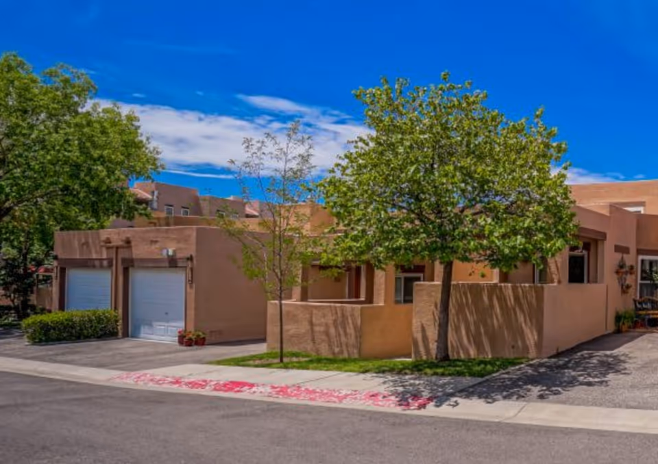 Exterior of a southwestern-style stucco residential building with two garage doors, courtyard walls, and trees along the sidewalk.