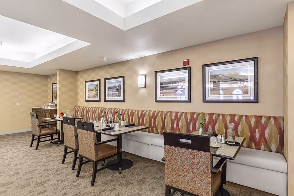 A dining area with tables and chairs arranged along a wall with patterned cushioned bench seating. The tables are set with cups, glasses, and small flower vases. The wall behind the seating features framed landscape artwork and a wall-mounted light fixture. The room has a beige color scheme with carpeted flooring and a recessed ceiling light.