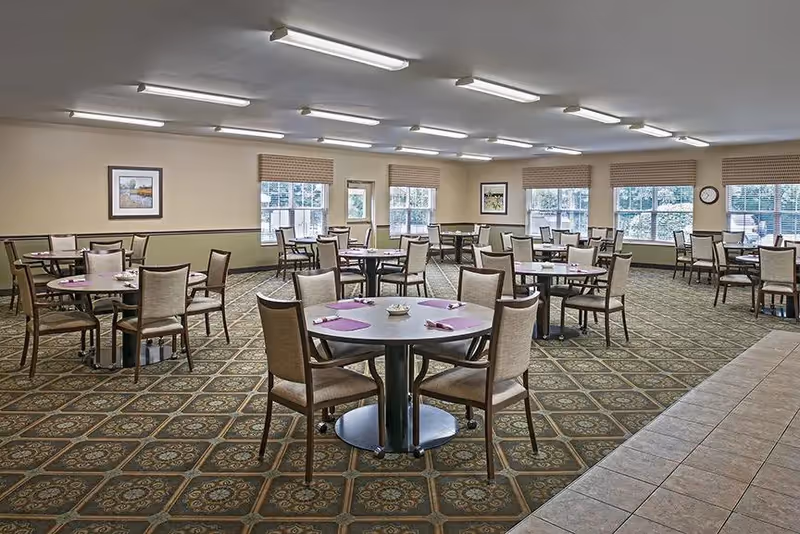 A spacious dining room with multiple round tables, each surrounded by four chairs. The tables are set with purple placemats and napkins. The room has large windows with beige blinds, patterned carpet flooring, and framed artwork on the walls. Fluorescent lights illuminate the ceiling.