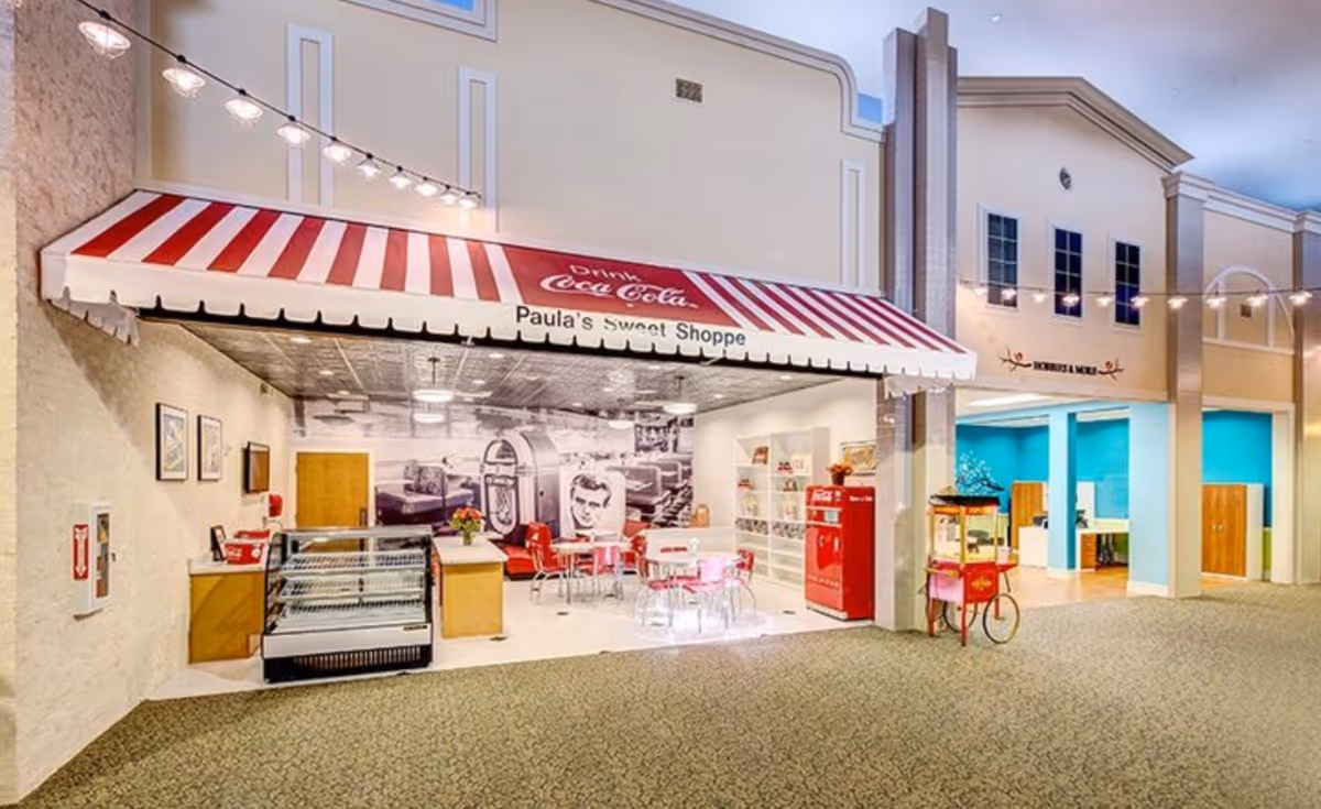 Indoor area designed to look like a vintage sweet shop with a red and white striped awning labeled 'Paula's Sweet Shoppe' and 'Drink Coca-Cola'. Inside, there are red and pink chairs and tables, a red vintage Coca-Cola vending machine, a popcorn cart, and a display case. The background features a large black and white mural of a retro diner scene. Adjacent to the sweet shop is an open area with blue walls and wooden cabinets.