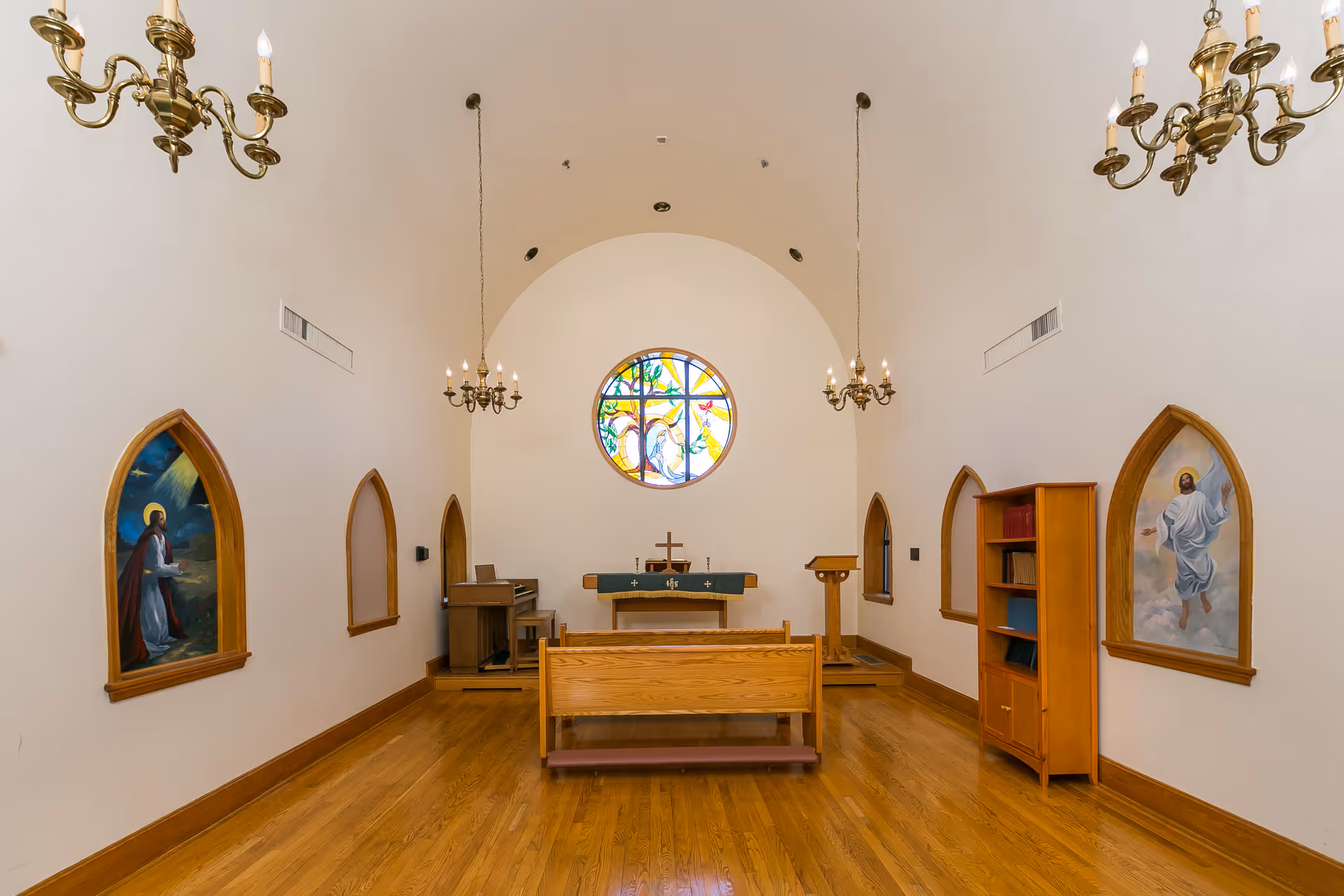 Interior chapel with wooden pews facing an altar beneath a round stained-glass window, chandeliers, and religious paintings on the walls.
