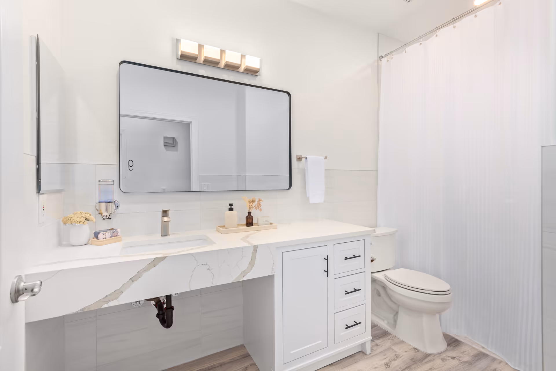 A clean, modern bathroom with a white marble countertop sink, a large rectangular mirror with black frame, a white toilet, and a white shower curtain. The bathroom has light-colored walls and wood-style flooring, with a towel hanging on a rack next to the mirror.