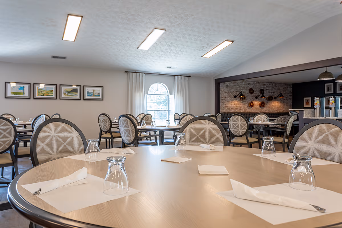 Dining room with round tables set with napkins and upside-down glasses, patterned chairs, and a window letting in natural light.