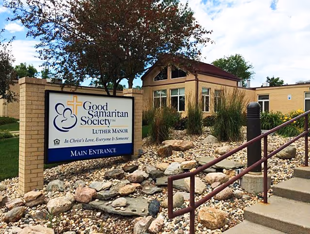Exterior view of Good Samaritan Society - Luther Manor showing the main entrance sign surrounded by rocks and landscaping with a building and trees in the background under a partly cloudy sky.
