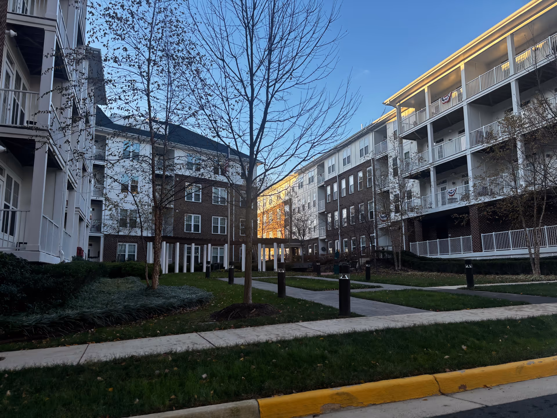 Exterior view of a senior living facility named Overture Fair Ridge, showing multi-story buildings with balconies, leafless trees, green lawns, and paved walkways under a clear blue sky.