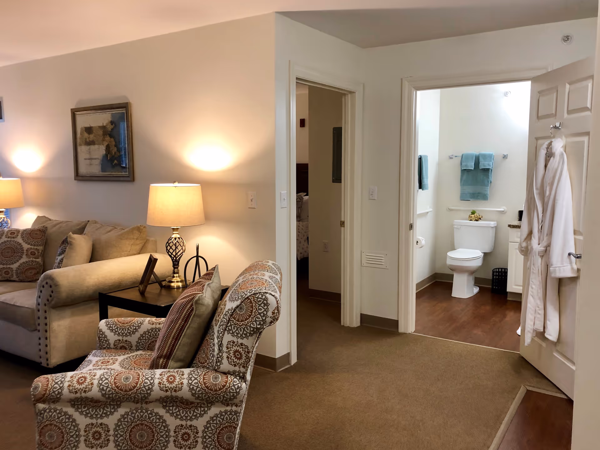 Interior view of a senior living facility showing a cozy living room with a beige sofa, patterned armchair, side table with a lamp, and a framed picture on the wall. Two open doorways lead to a bedroom and a bathroom with a toilet, towels, and a bathrobe hanging on the door.