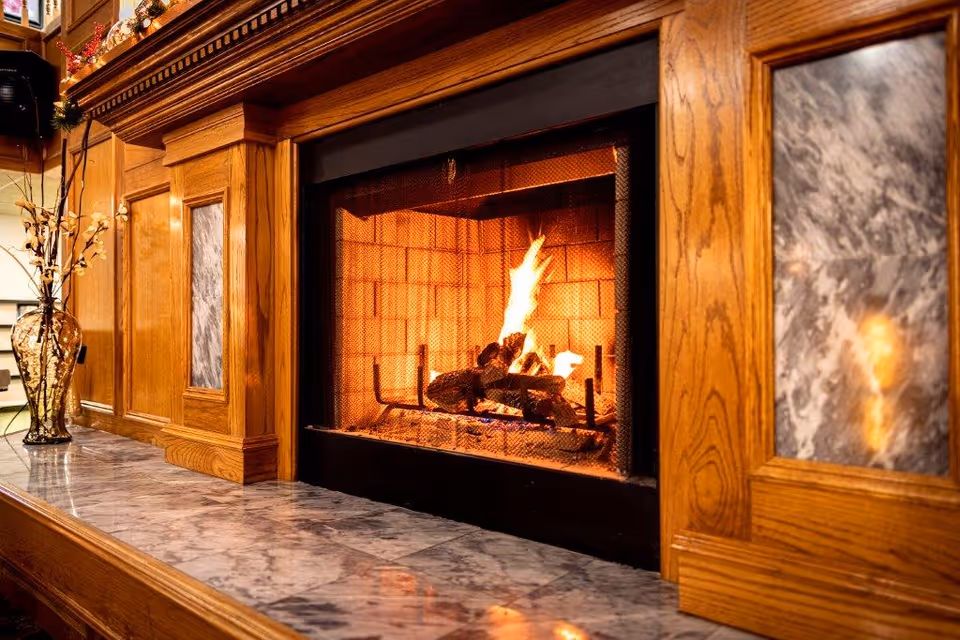 A cozy fireplace with a wooden mantel and marble accents. The fire is burning brightly with logs inside the fireplace. A decorative vase with branches is placed on the marble surface next to the fireplace.