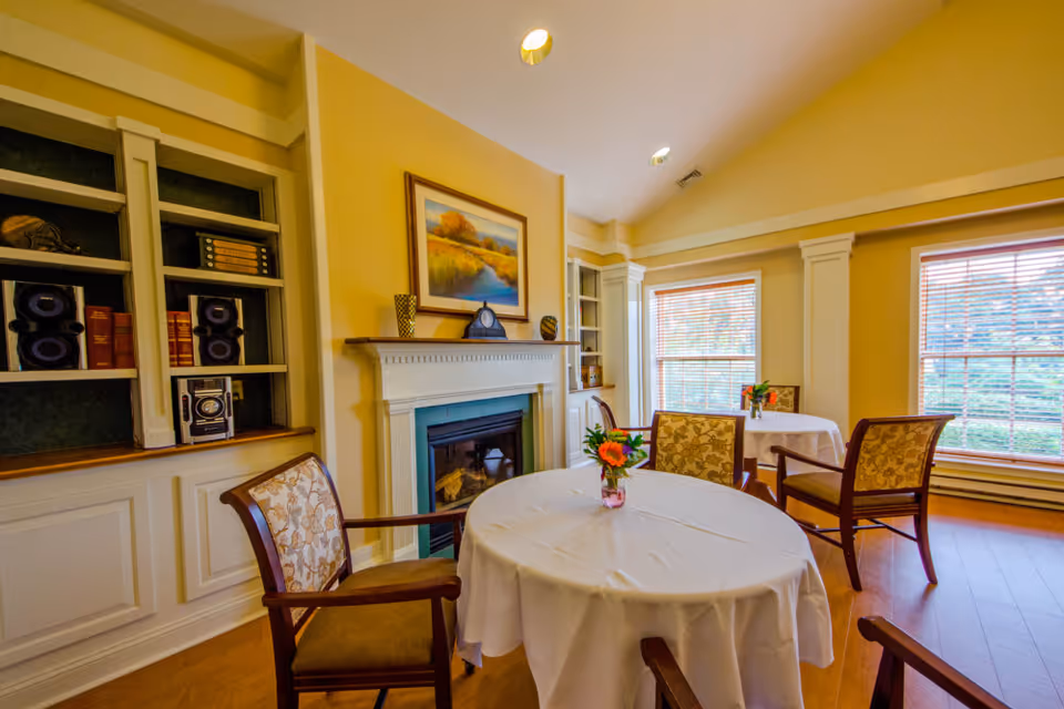 A cozy dining area with round tables covered in white tablecloths, each adorned with a small vase of flowers. The room features wooden chairs with patterned upholstery, large windows with blinds letting in natural light, a fireplace with a painting above it, and built-in shelves with books and decorative items.