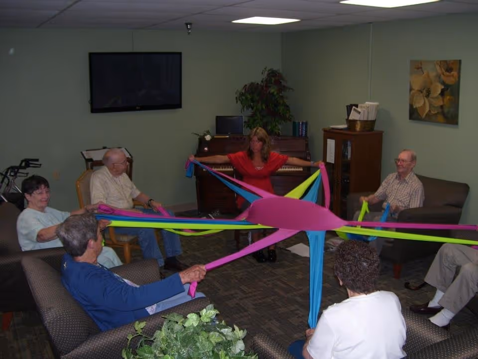A group of elderly residents and a staff member sit in a lounge holding colorful fabric bands stretched across a circle for a group activity.