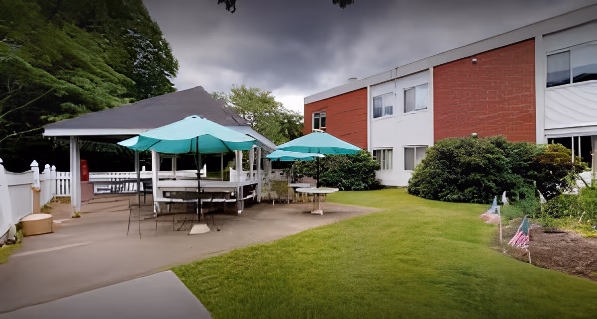 Outdoor courtyard with covered picnic tables and teal umbrellas next to a two-story brick-and-white building.