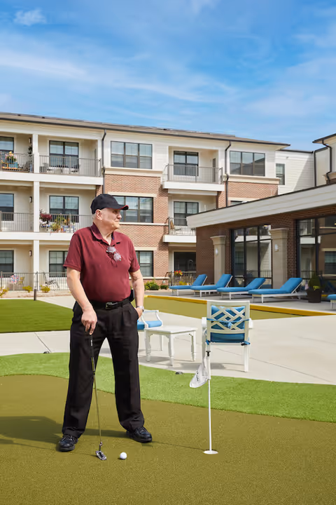 An elderly man wearing a maroon polo shirt, black pants, and a black cap stands on a putting green holding a golf club. Behind him is a modern senior living facility with balconies, lounge chairs, and a clear blue sky.