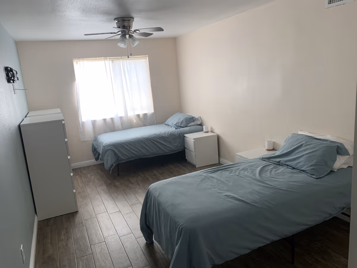 A bright bedroom with two single beds dressed in blue-gray linens, matching nightstands, a dresser, wood-look flooring and a ceiling fan.