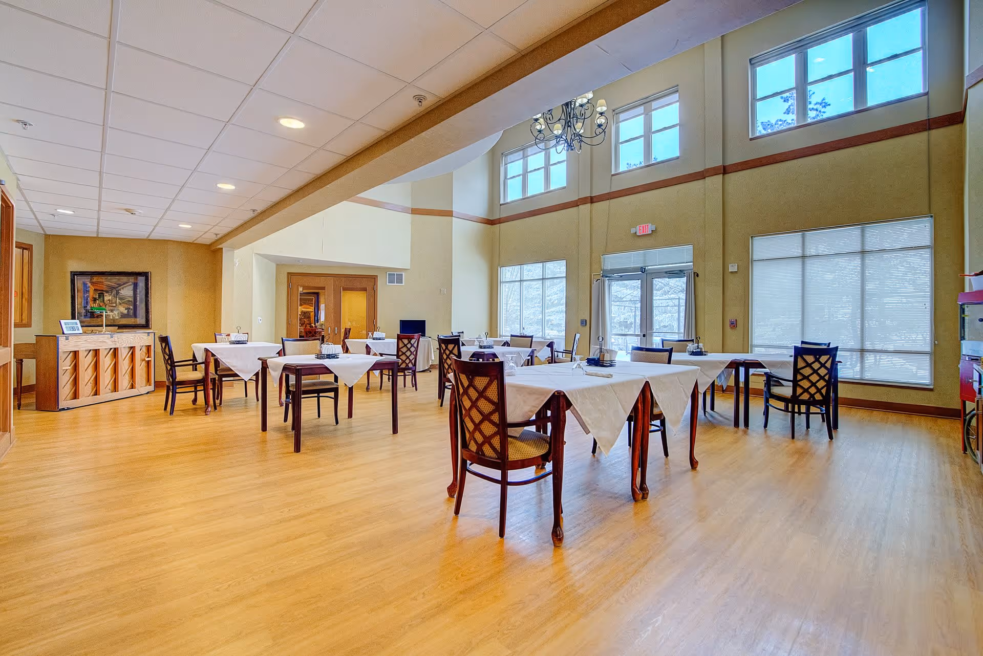 A spacious dining room with several tables covered with white tablecloths and wooden chairs arranged around them. The room has large windows letting in natural light, a chandelier hanging from the high ceiling, and a piano against one wall. The floor is wooden, and the walls are painted in warm tones.