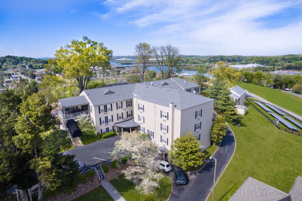 Aerial view of a multi-story senior living facility building surrounded by trees and greenery, with a parking area and a driveway. In the background, there is a river, a bridge, and a small town with houses and other buildings under a partly cloudy blue sky.
