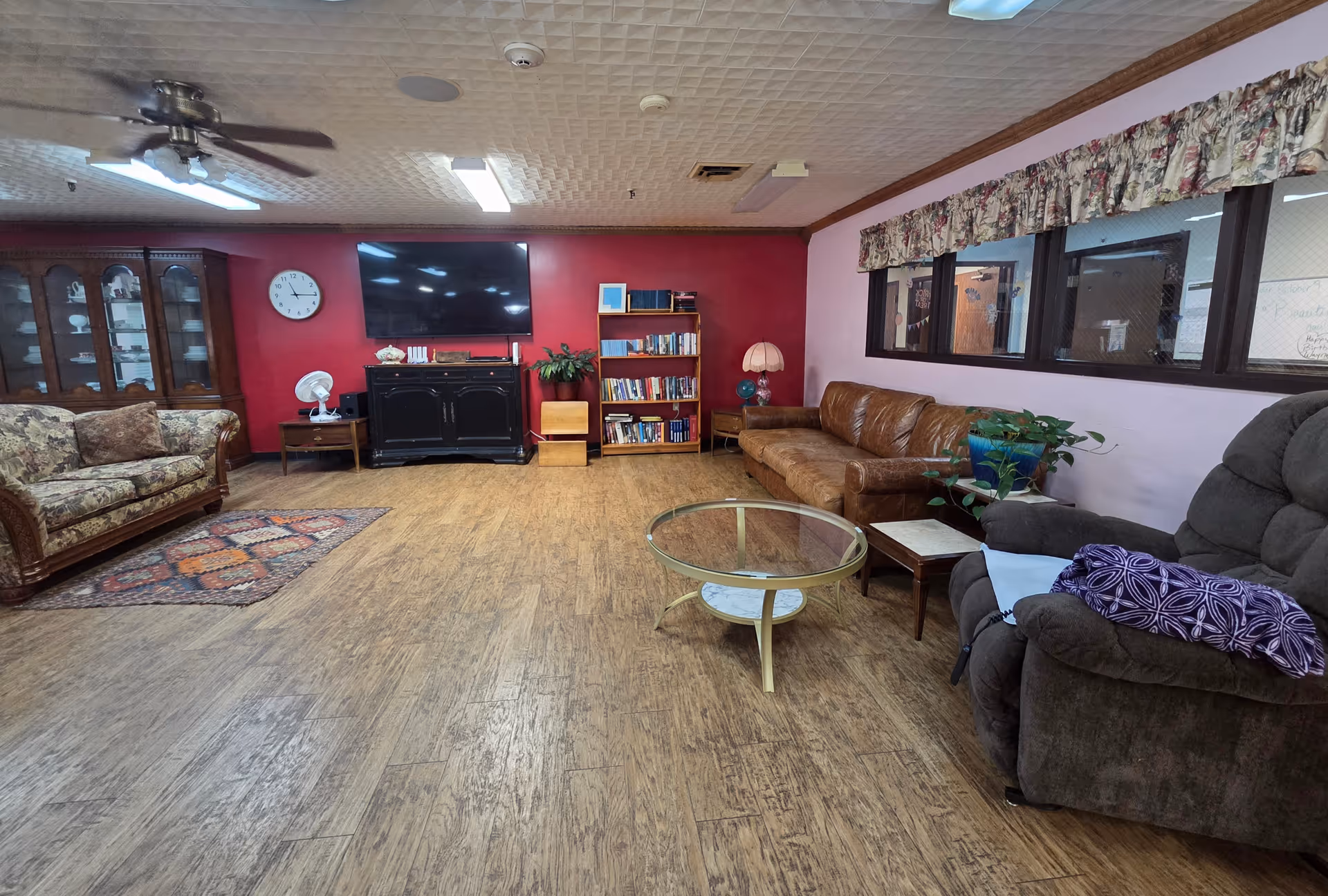 A cozy nursing home common room with sofas, a recliner, a glass coffee table, a TV mounted on a red accent wall, a bookshelf, and wood-look flooring.