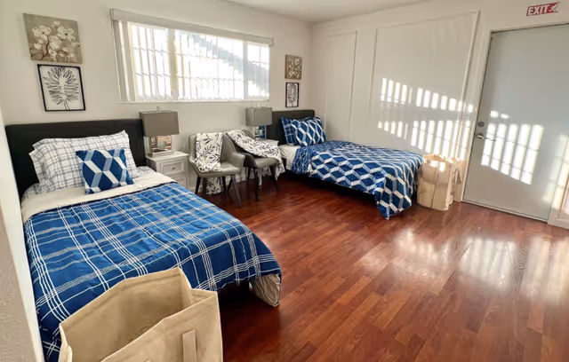 Sunlit room with two twin beds dressed in blue patterned bedding, chairs, and a door with an exit sign.