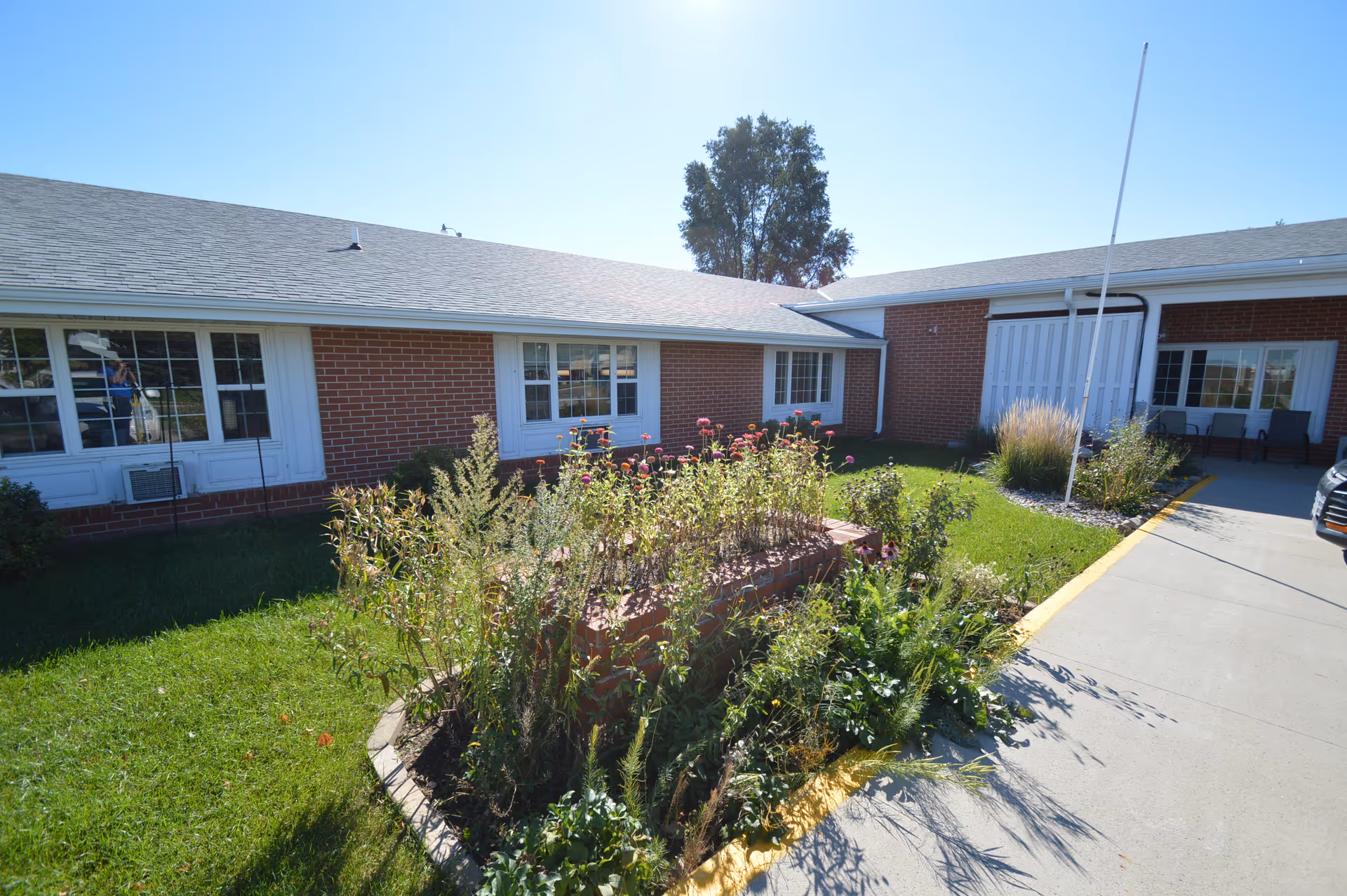 Front exterior of a one-story brick senior living building with windows, a raised flowerbed, a flagpole and a driveway entrance.