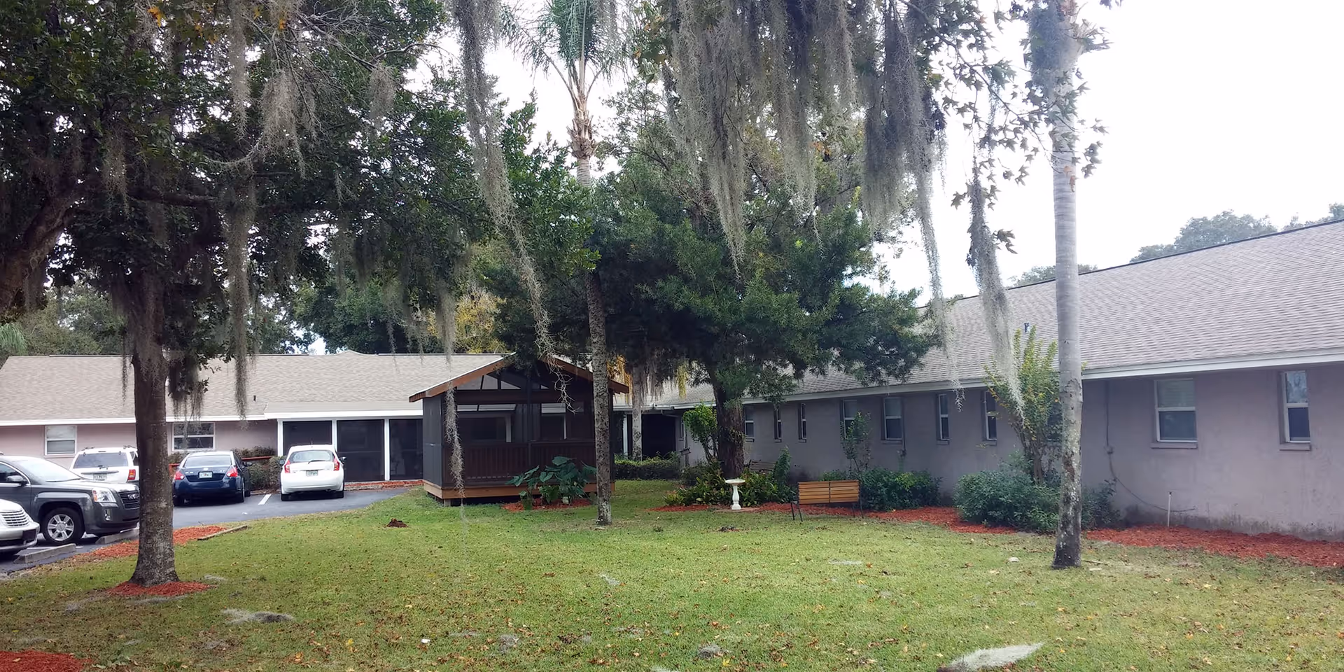 Outdoor view of a senior living facility with a grassy courtyard area surrounded by single-story buildings. There are several trees with hanging moss, a wooden bench, a birdbath, and a small covered pavilion. Several parked cars are visible on the left side near the building.