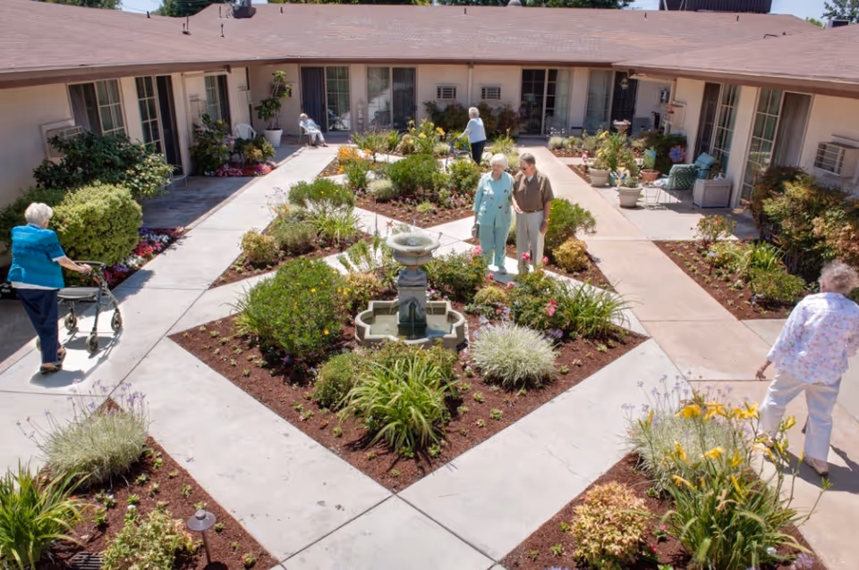 Outdoor courtyard garden area at a senior living facility with paved walkways, flower beds, and a central water fountain. Several elderly people are walking or sitting around the garden, enjoying the sunny day. The courtyard is surrounded by single-story building rooms with large windows and doors.