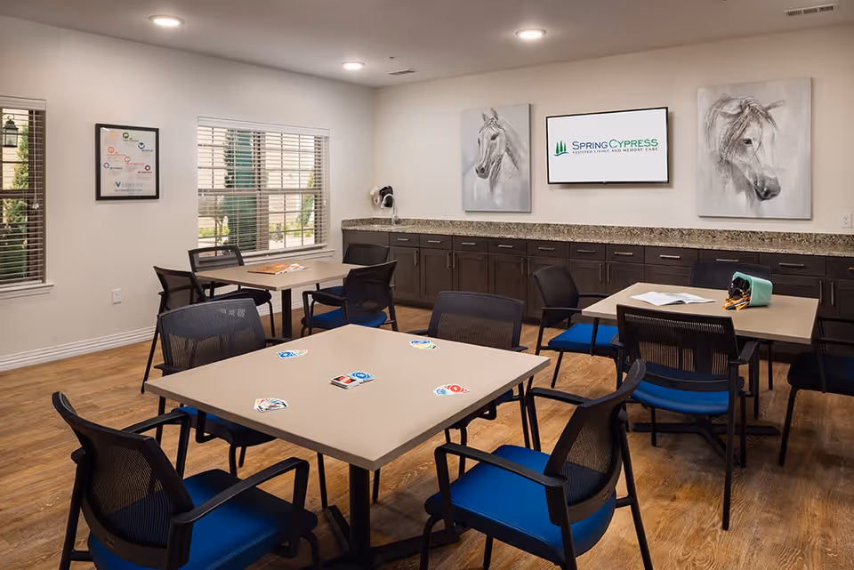 A well-lit common room with several tables and chairs arranged for group activities. The tables have playing cards and a book on them. The room features large windows with blinds, dark wood cabinets with a granite countertop along one wall, and three framed artworks including two horse paintings and a screen displaying the Spring Cypress logo.