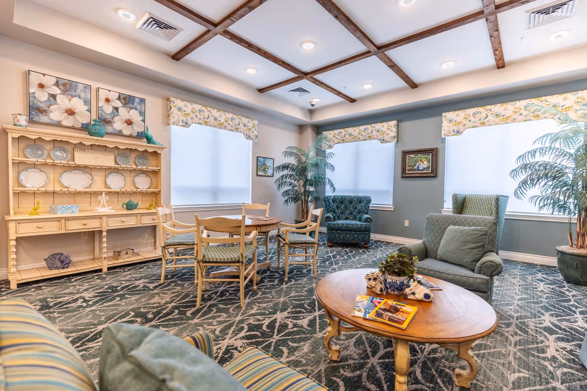 A cozy living room area in an assisted living facility featuring a round wooden coffee table with magazines and a plant, several upholstered armchairs, a wooden dining table with chairs, a decorative hutch displaying plates, floral curtains on windows, and potted plants in the corners.
