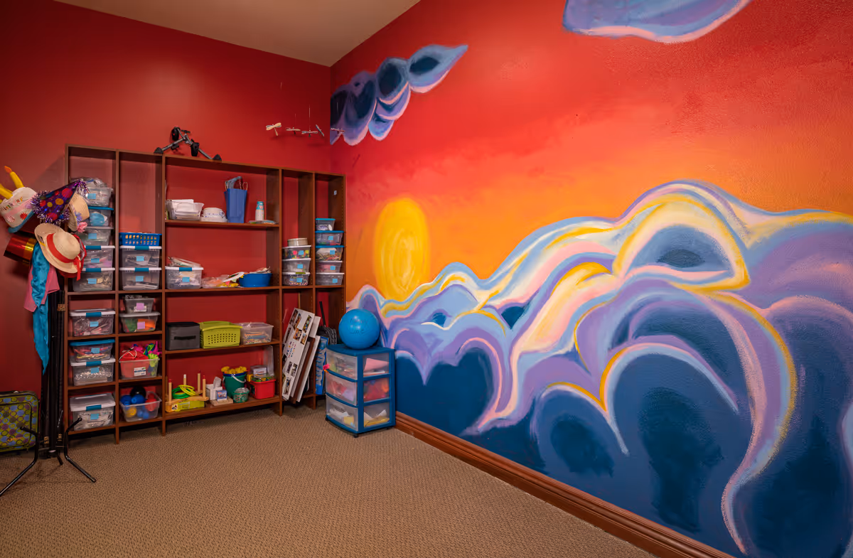 Colorful activity room with a sun-and-cloud mural on a red wall and shelving filled with craft supplies and bins.