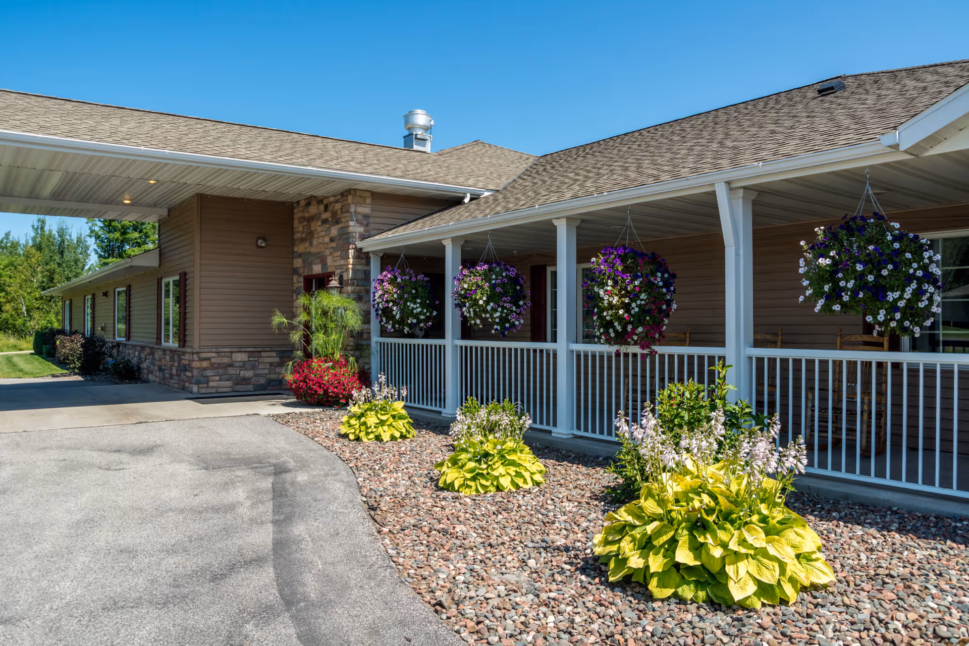 Front entrance of a senior living building with a covered porch, hanging flower baskets, and landscaped rock beds.