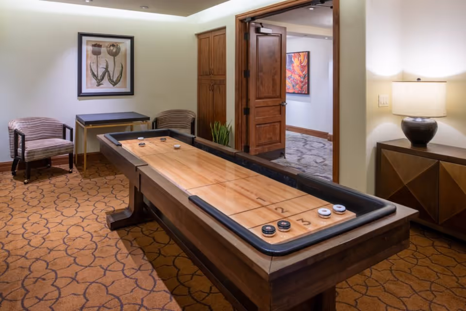 A cozy indoor game room with a shuffleboard table in the center. The room features patterned carpet, two striped armchairs, a small table, a wooden cabinet with a lamp, and framed artwork on the walls. A wooden door is open, revealing a hallway with additional artwork.