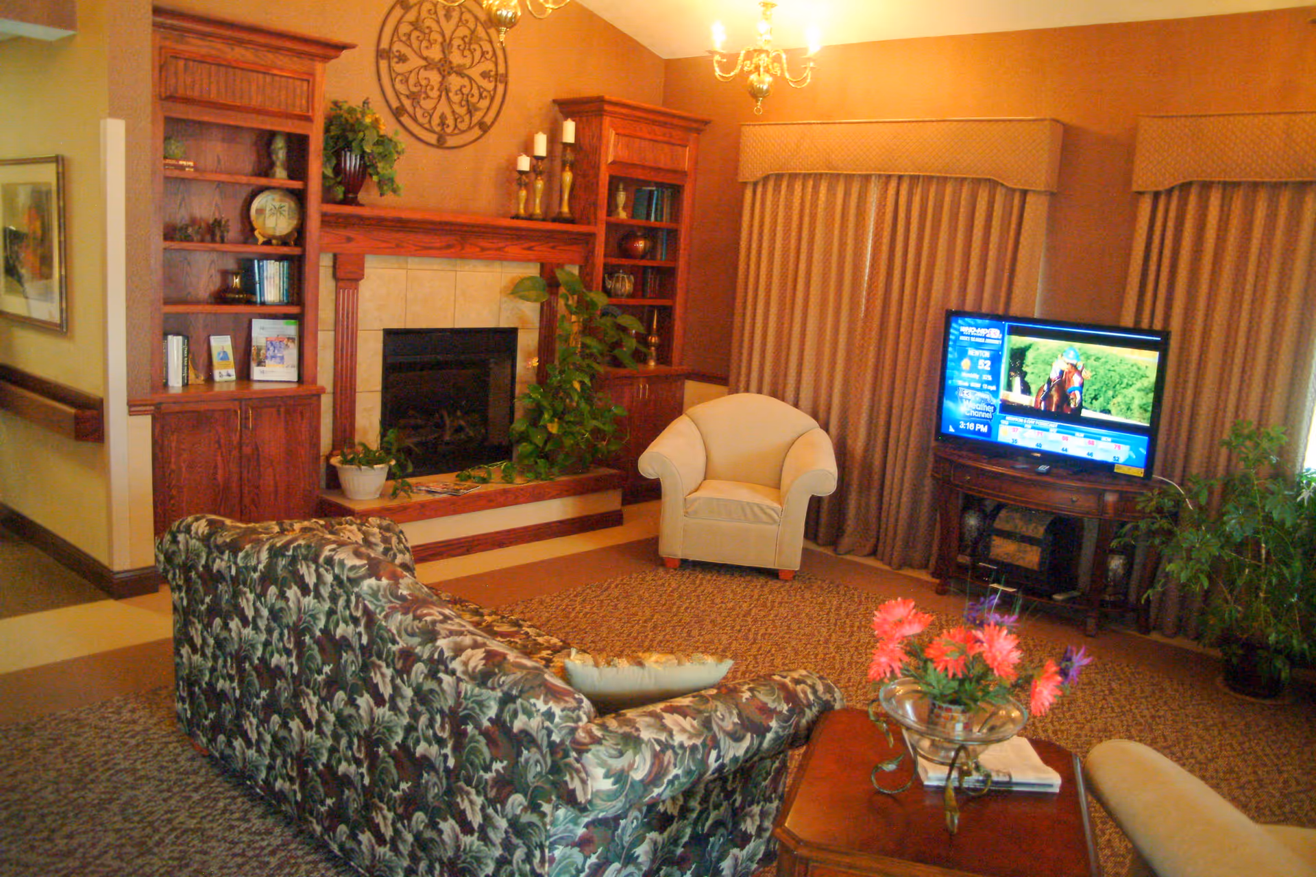 Cozy living room with a floral sofa, armchair, fireplace flanked by built-in bookshelves, TV on a stand, and a coffee table with flowers.