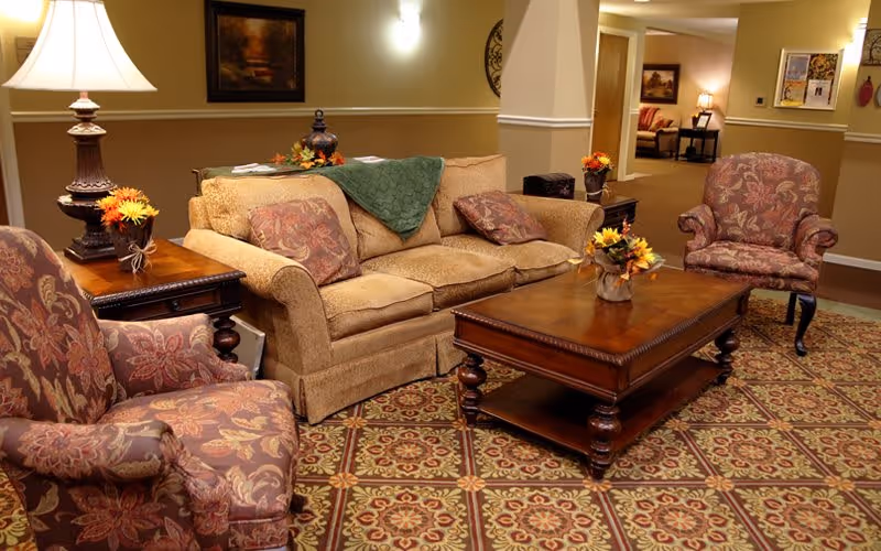 A cozy living room area with a beige sofa adorned with patterned cushions and a green throw blanket. Two upholstered armchairs with floral patterns flank a wooden coffee table decorated with a small flower arrangement. The room features warm lighting from a table lamp and wall sconces, with framed artwork on the walls and a patterned carpet covering the floor.