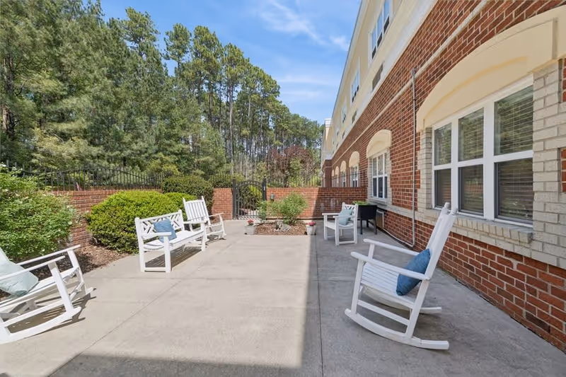 Outdoor patio area at Fox Hollow Senior Living Community with white rocking chairs and benches arranged on a concrete surface next to a red brick building. The patio is bordered by green bushes, a brick wall, and tall trees under a blue sky with some clouds.
