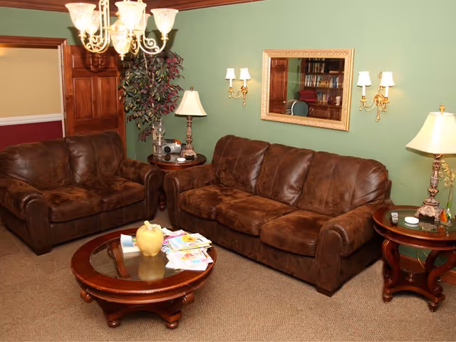 A cozy living room with two brown leather sofas, a round wooden coffee table with magazines and a decorative item on it, two wooden side tables each with a lamp, a green wall with a large framed mirror, two wall-mounted sconces with lampshades, a chandelier hanging from the ceiling, and a potted plant in the corner.