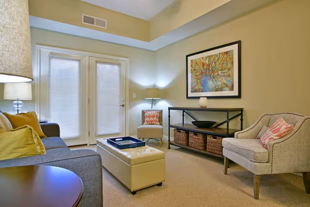 Well-lit living room with a sofa, ottoman, two armchairs, a console table with baskets, framed artwork, lamps, and French doors with blinds.
