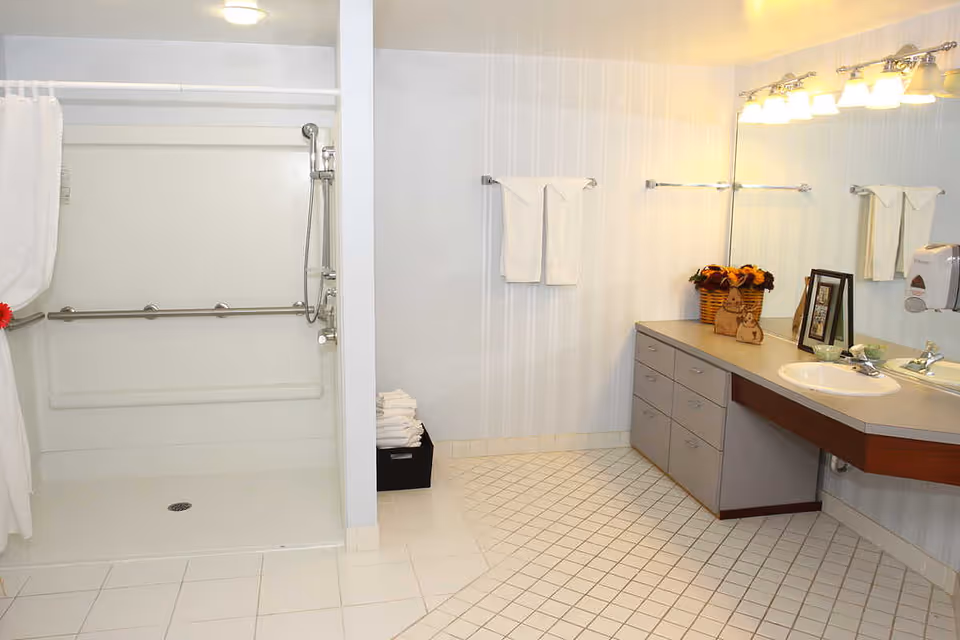 Accessible senior bathroom featuring a roll-in shower with grab bars, a long vanity and mirror, and neatly folded towels.