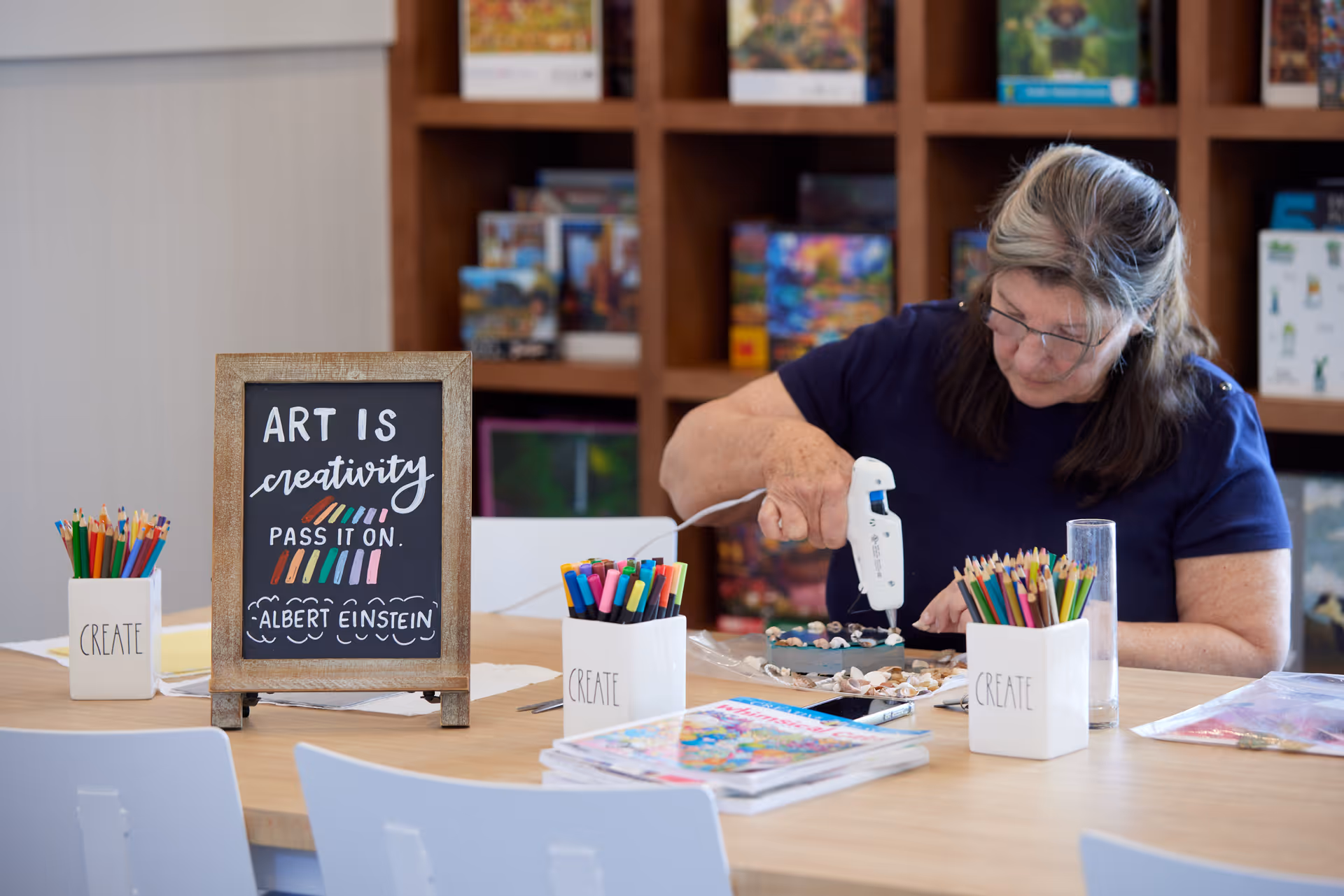 An elderly woman using a hot glue gun to create an art project at a table with colored pencils, markers, and magazines. A framed chalkboard sign on the table reads, 'Art is creativity, pass it on. Albert Einstein.' Shelves with puzzles and games are visible in the background.