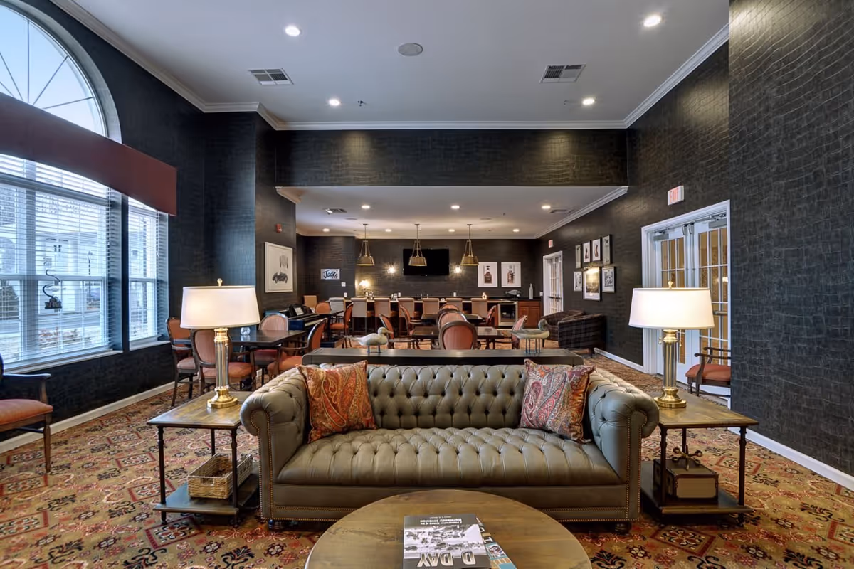 A spacious senior living common area with a green tufted sofa adorned with patterned pillows in the foreground. Two side tables with lamps flank the sofa. Behind the sofa are multiple tables and chairs arranged for dining or socializing. The room features dark textured walls, a large arched window with blinds on the left, and framed artwork on the walls. Pendant lights hang over a counter area at the back, and a television is mounted on the far wall.