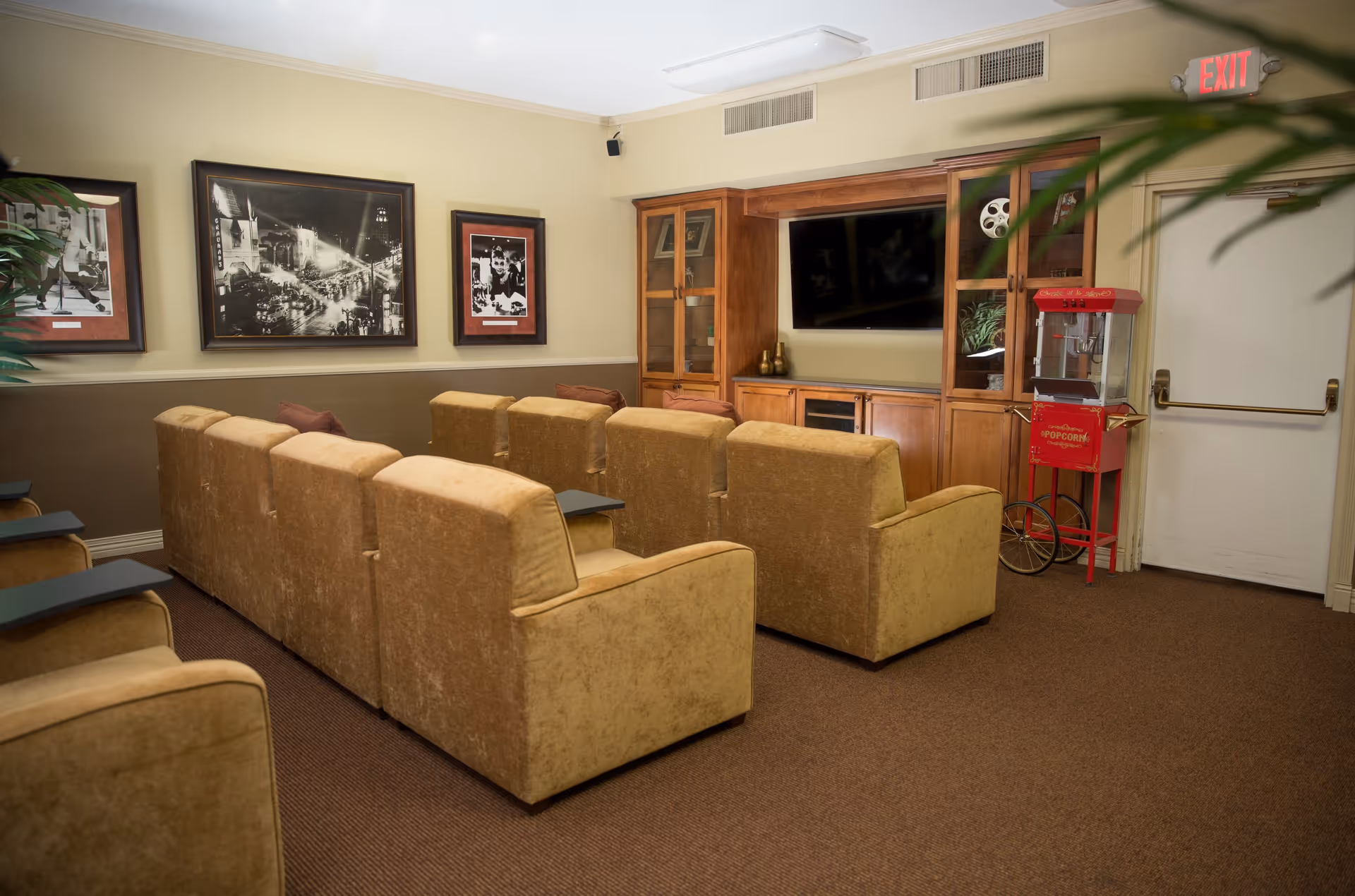 A cozy media room with two rows of beige upholstered armchairs facing a large flat-screen TV mounted on a wooden cabinet. The room has framed black and white photographs on the wall, a red vintage-style popcorn machine near an exit door, and a potted plant partially visible in the foreground.