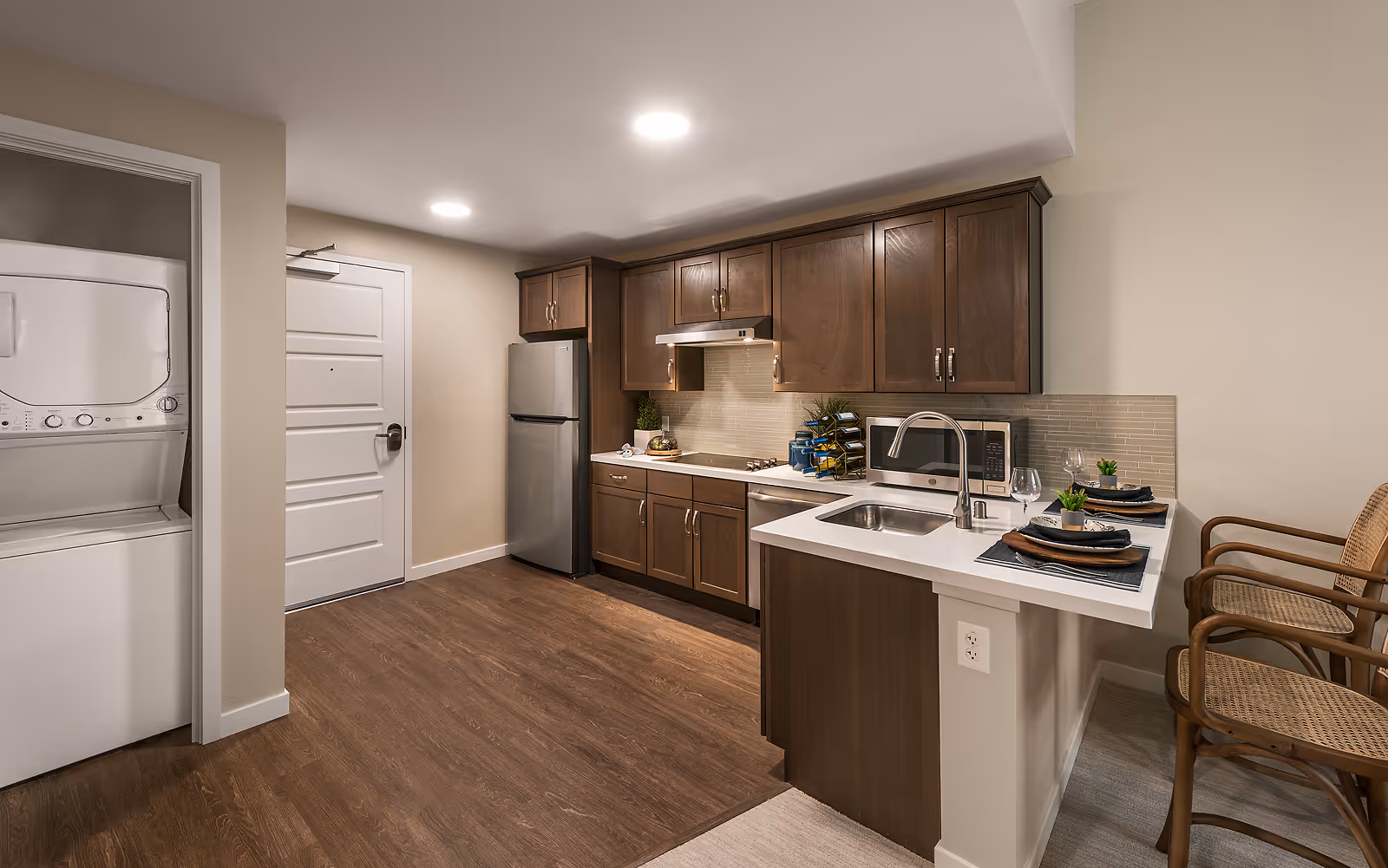 Modern kitchen area with dark wooden cabinets, stainless steel refrigerator, microwave, and a sink with a faucet on a white countertop. Two wooden chairs with woven seats are placed at the counter set with plates, napkins, and wine glasses. To the left, there is a stacked washer and dryer unit next to a white door. The floor is a combination of wood and carpet.